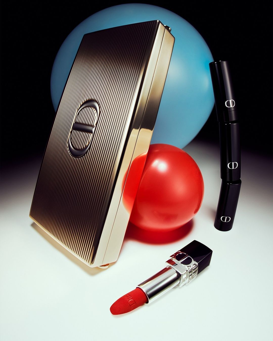 Cosmetic still life: Luxurious gold Dior compact, black lipsticks, and red lipstick against blue and red orbs on white surface.