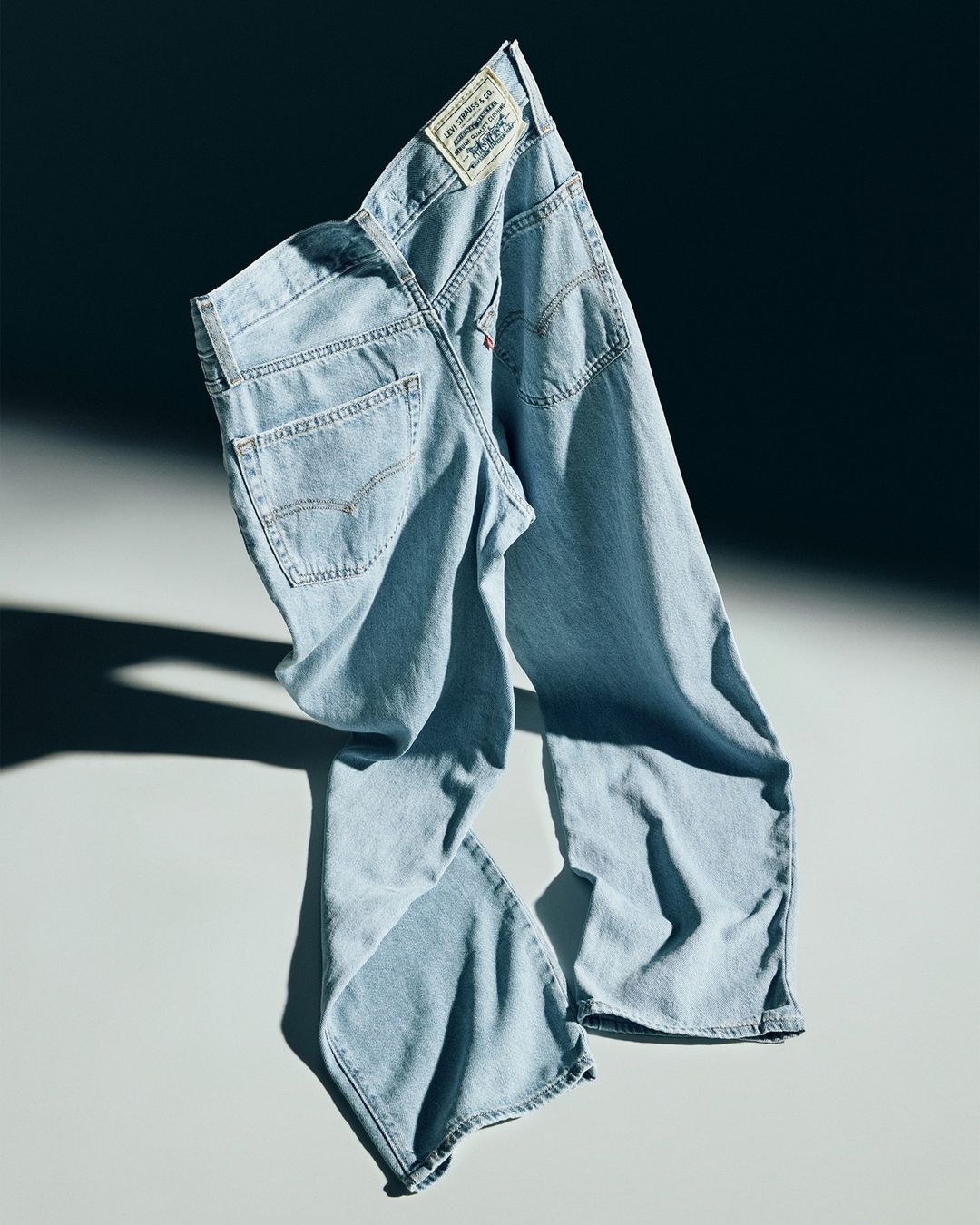 Light wash denim jeans on a light grey background in studio lighting. Dark shadow is cast beside the garment. Brand label visible.