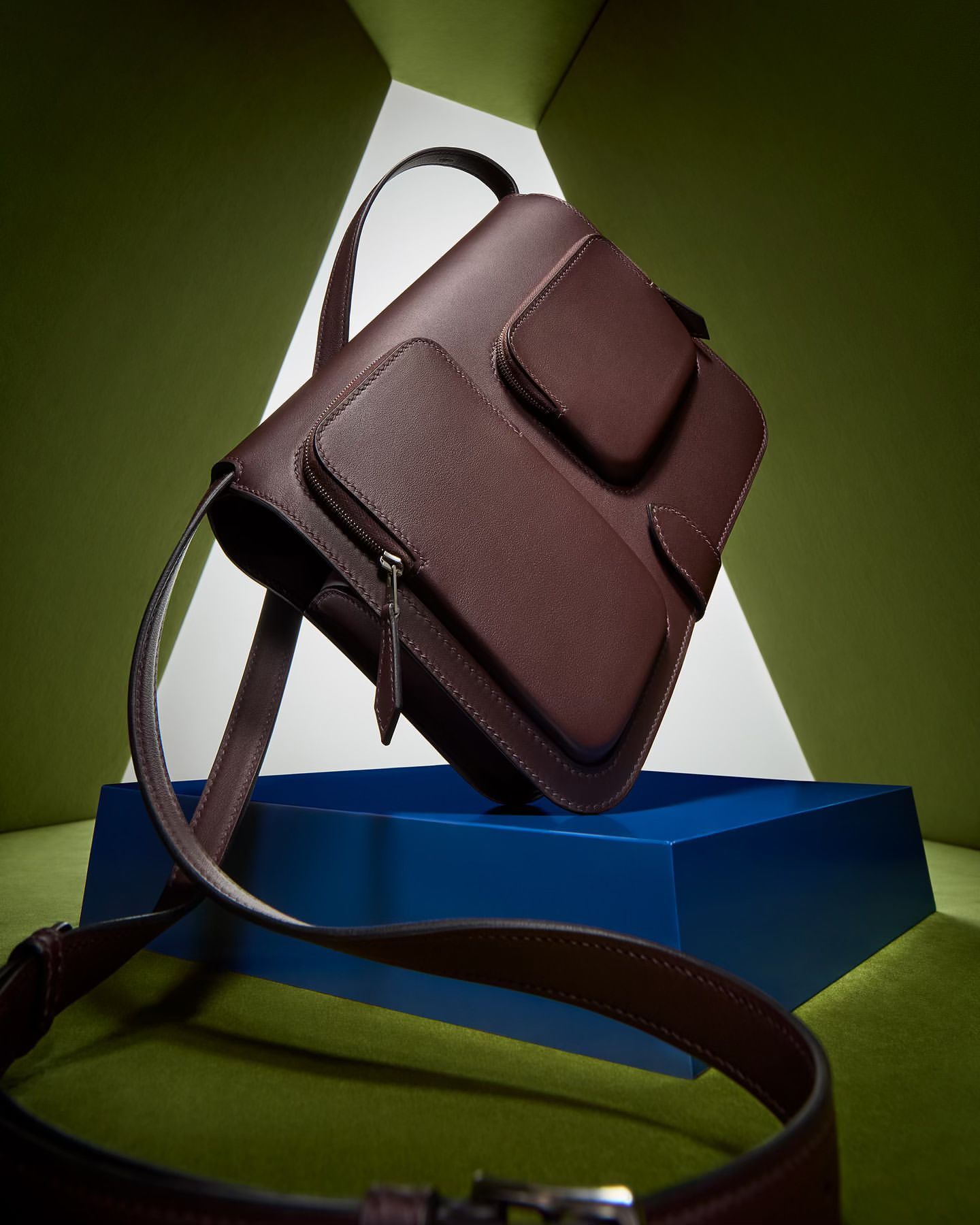Studio shot of a maroon leather satchel handbag with front pockets on a dark blue platform, set against a geometric green backdrop.