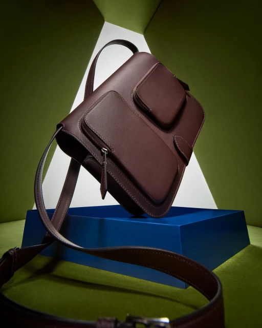 Studio shot of a maroon leather satchel handbag with front pockets on a dark blue platform, set against a geometric green backdrop.