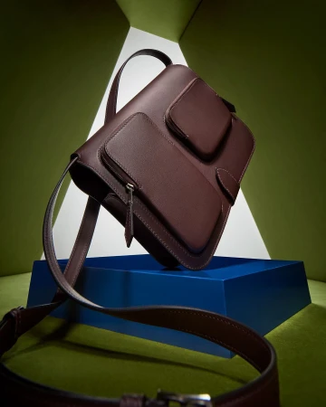 Studio shot of a maroon leather satchel handbag with front pockets on a dark blue platform, set against a geometric green backdrop.