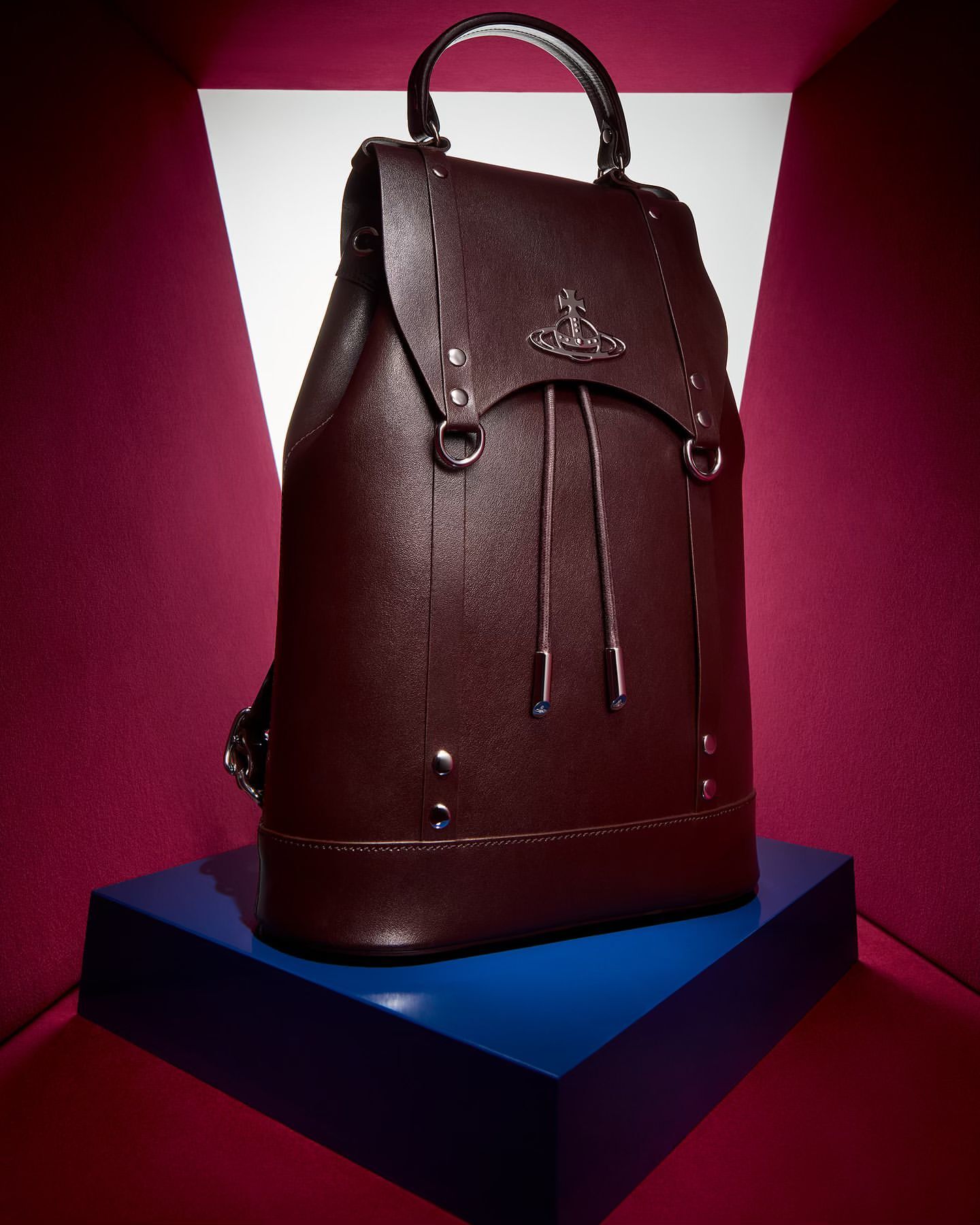 Burgundy leather backpack with a rounded top handle and signature logo detail, displayed against a backdrop of vibrant fuchsia and white.