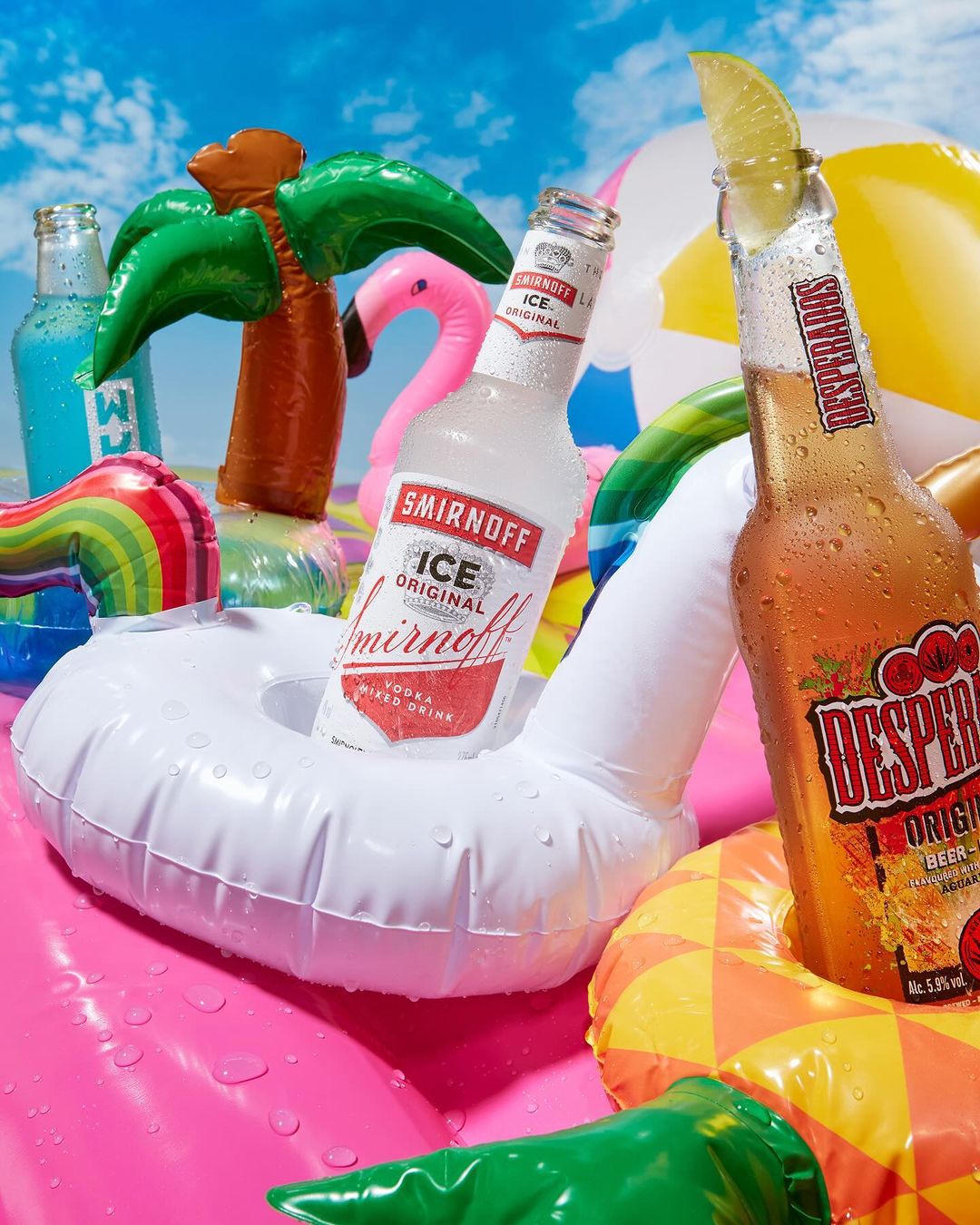 Two bottles of alcoholic beverages in floatation devices, surrounded by inflatable pool toys, set against a blue sky with light clouds.