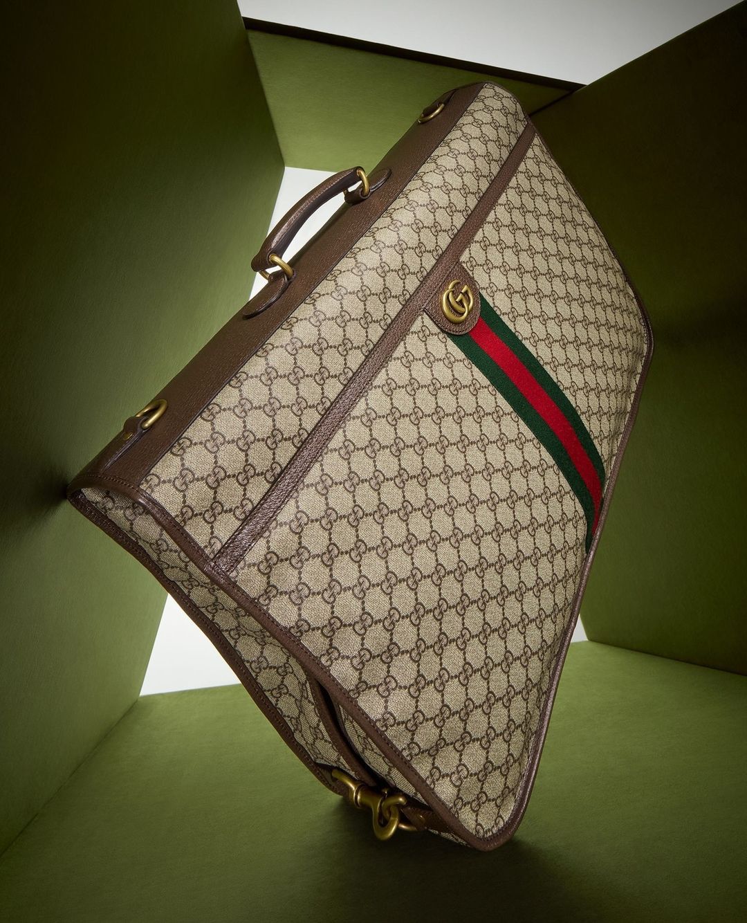Gucci garment bag featuring the brand's monogram pattern, brown leather trim, gold hardware, and green and red stripe, shot indoors.