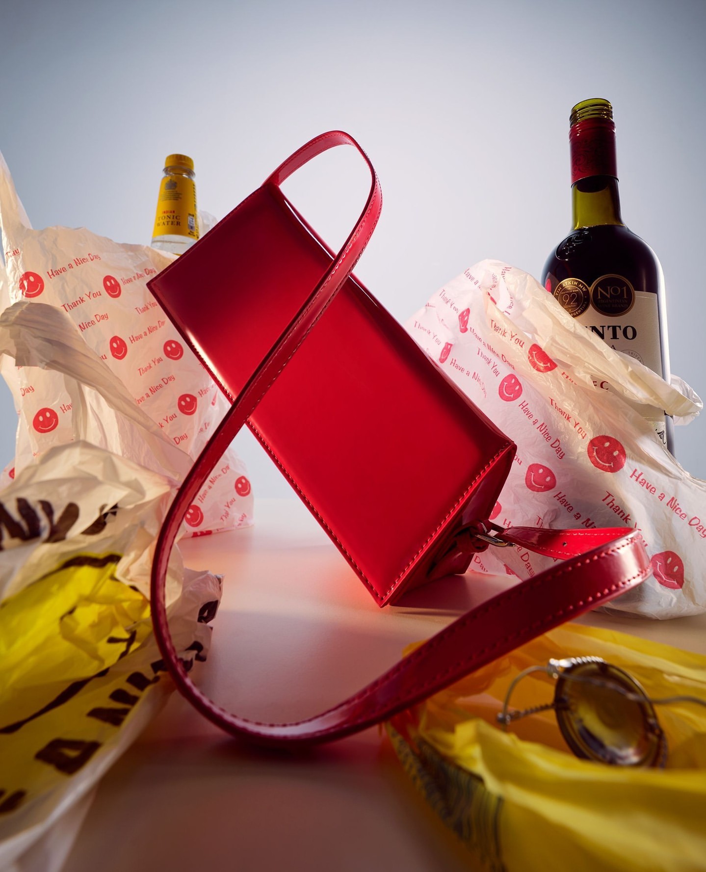 Red fashion purse surrounded by white and yellow plastic bags, wine bottle, and tonic bottle on white table in a product studio shot.