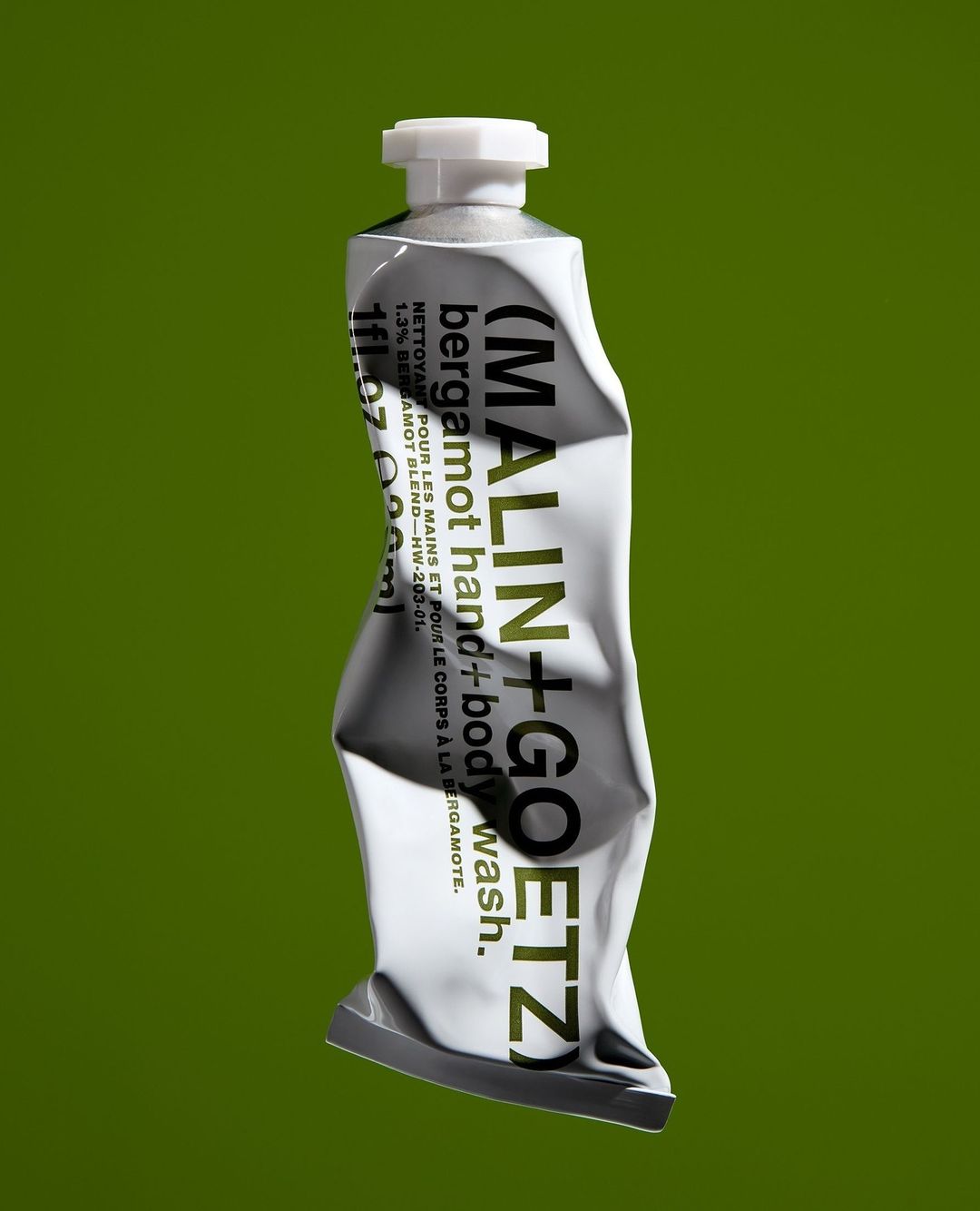 Close-up studio shot of a slightly crumpled silver tube of Malin + Goetz bergamot hand and body wash against a solid green background.
