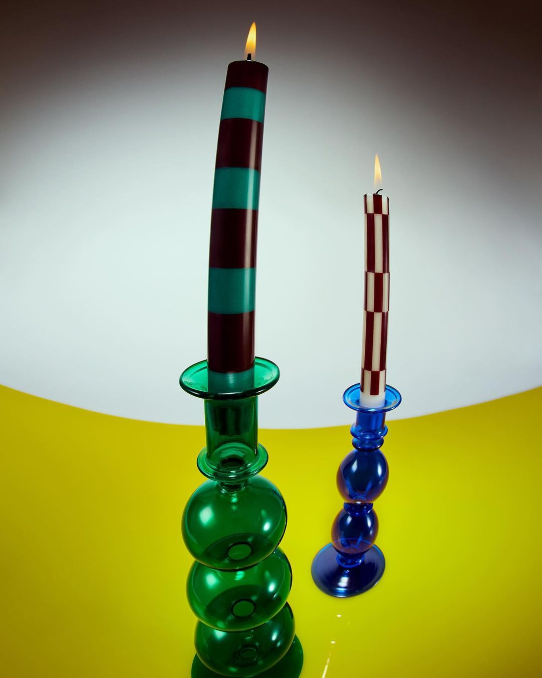 Two lit candles in decorative glass candle holders, one striped in brown and turquoise, the other checkered in brown and white.