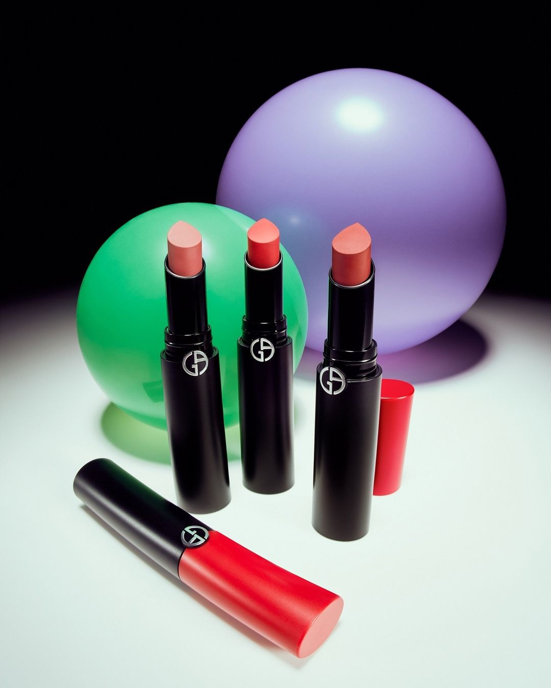 Cosmetics still life: three Giorgio Armani lipsticks stand among colorful balloons, set against a dark backdrop in a beauty product shot.