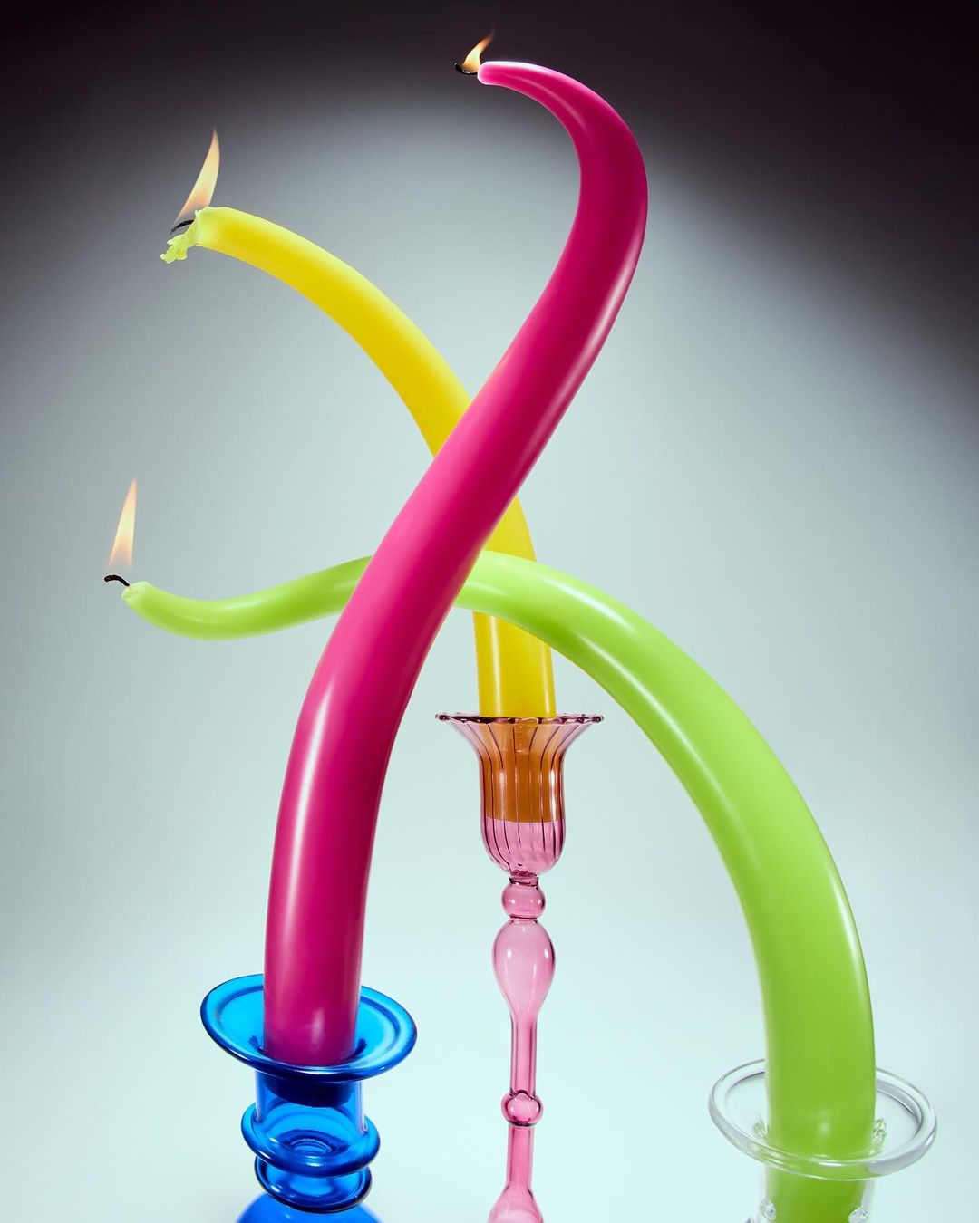 Colorful, bent candles in blue, pink, and clear candleholders. Each candle has a small, bright flame burning at the end.