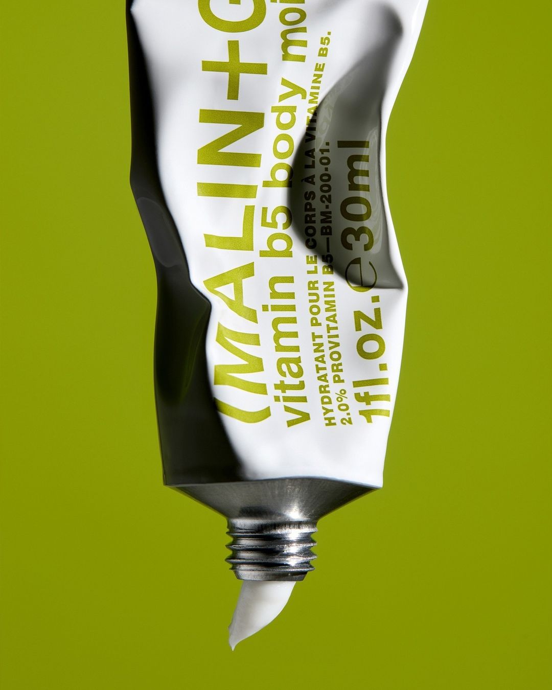 Close-up of a white tube of MALIN+GOETZ vitamin B5 body moisturizer against a green background, featuring product details and branding.