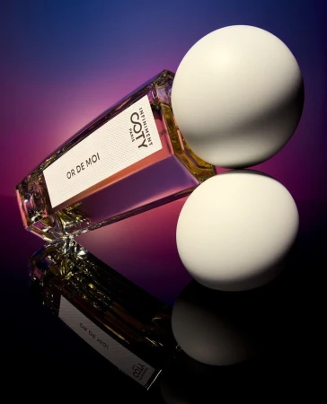Coty Or De Moi perfume bottle resting diagonally on a reflective surface alongside two smooth white spherical props against a gradient background.