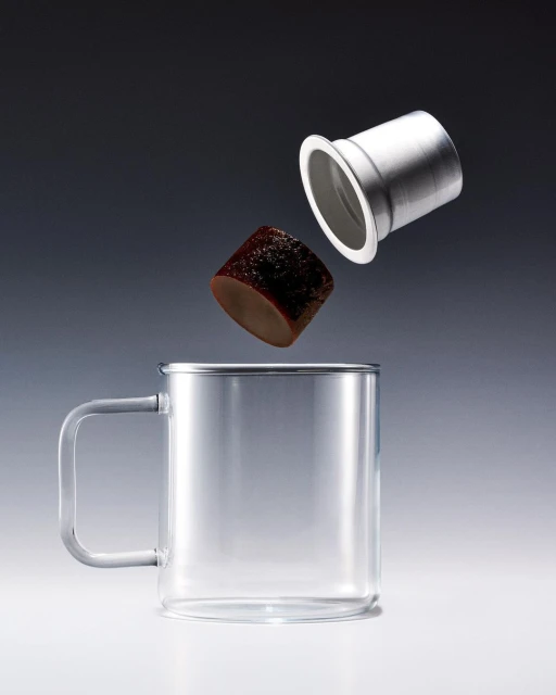 Studio shot of a clear mug, dark coffee cube, and a silver metal coffee filter on a gradient gray and white background. Food and beverage.