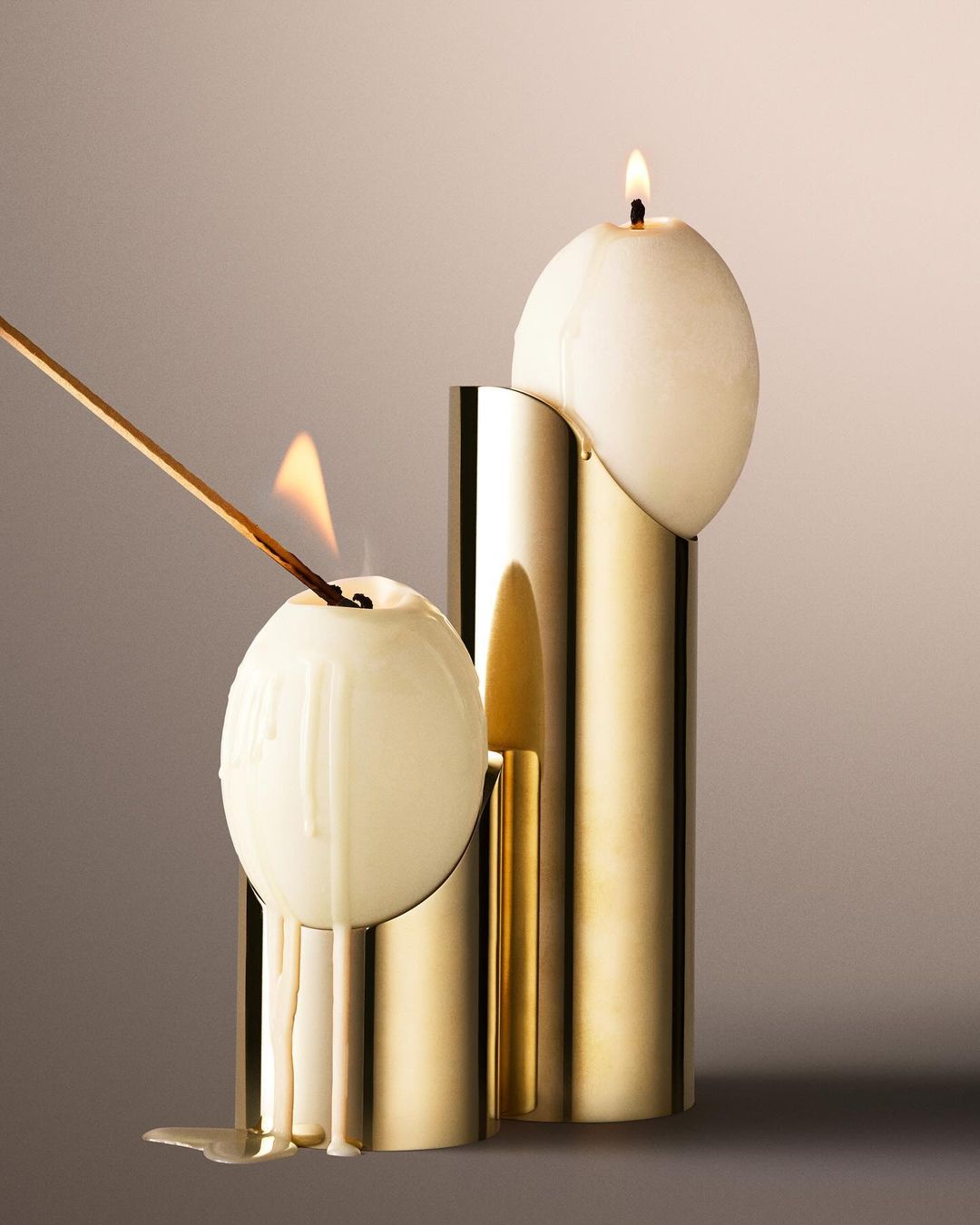Two sphere candles in gold tubular stands; one lit with flame and dripping wax after lighting with match stick in a studio setting.