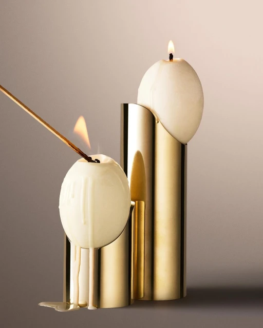 Two sphere candles in gold tubular stands; one lit with flame and dripping wax after lighting with match stick in a studio setting.