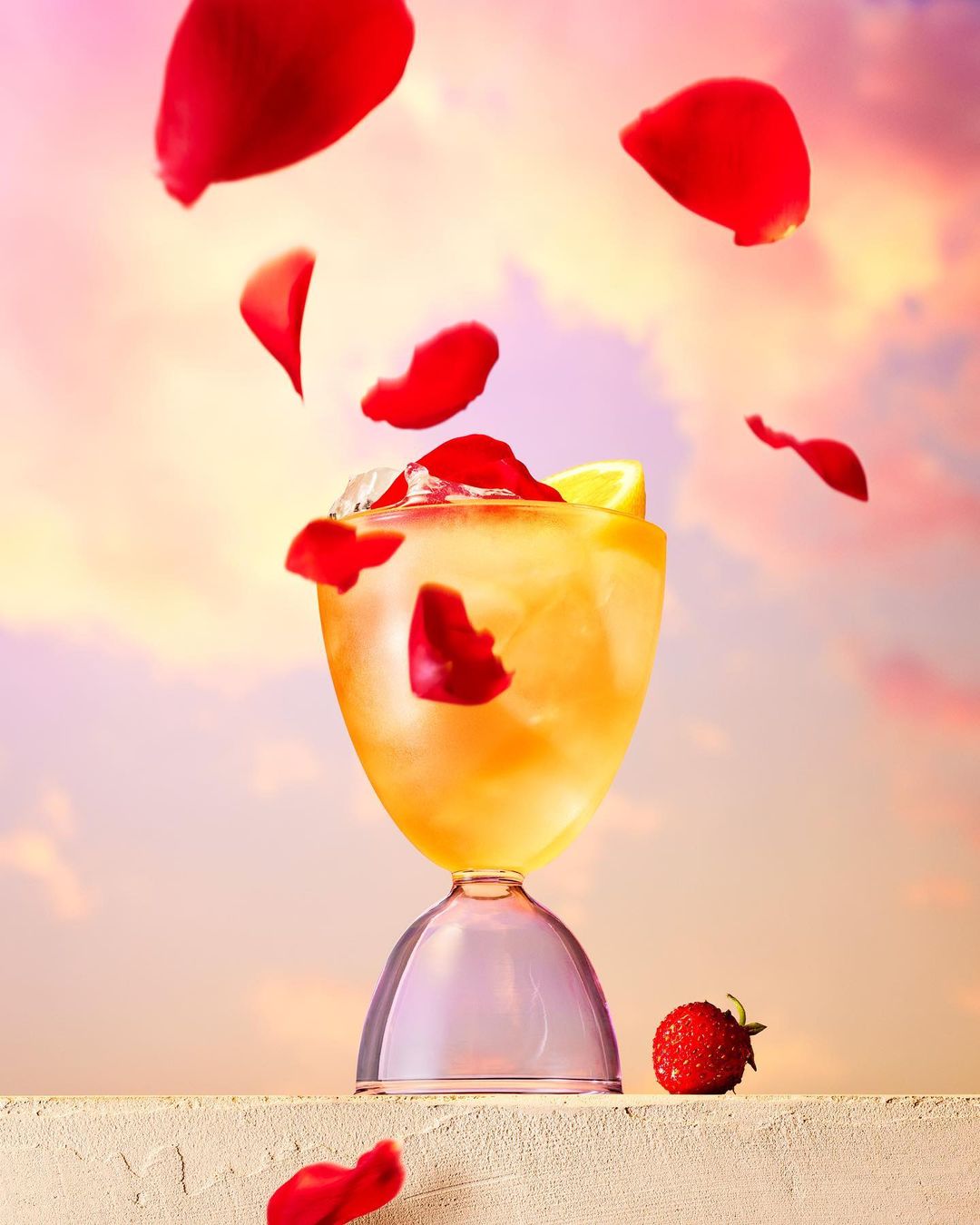 Elegant cocktail in glass with ice, lemon, strawberry, and falling rose petals against a pink sky, creating a refreshing and romantic visual.