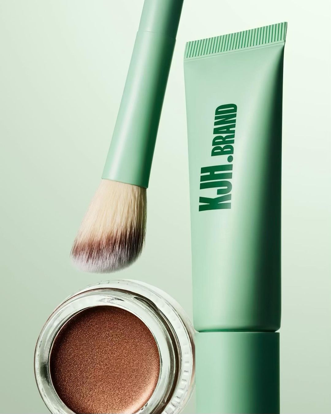 Cosmetic brush, tube of product with KJH.BRAND logo, and jar of bronzer on a light green reflective surface in a studio.