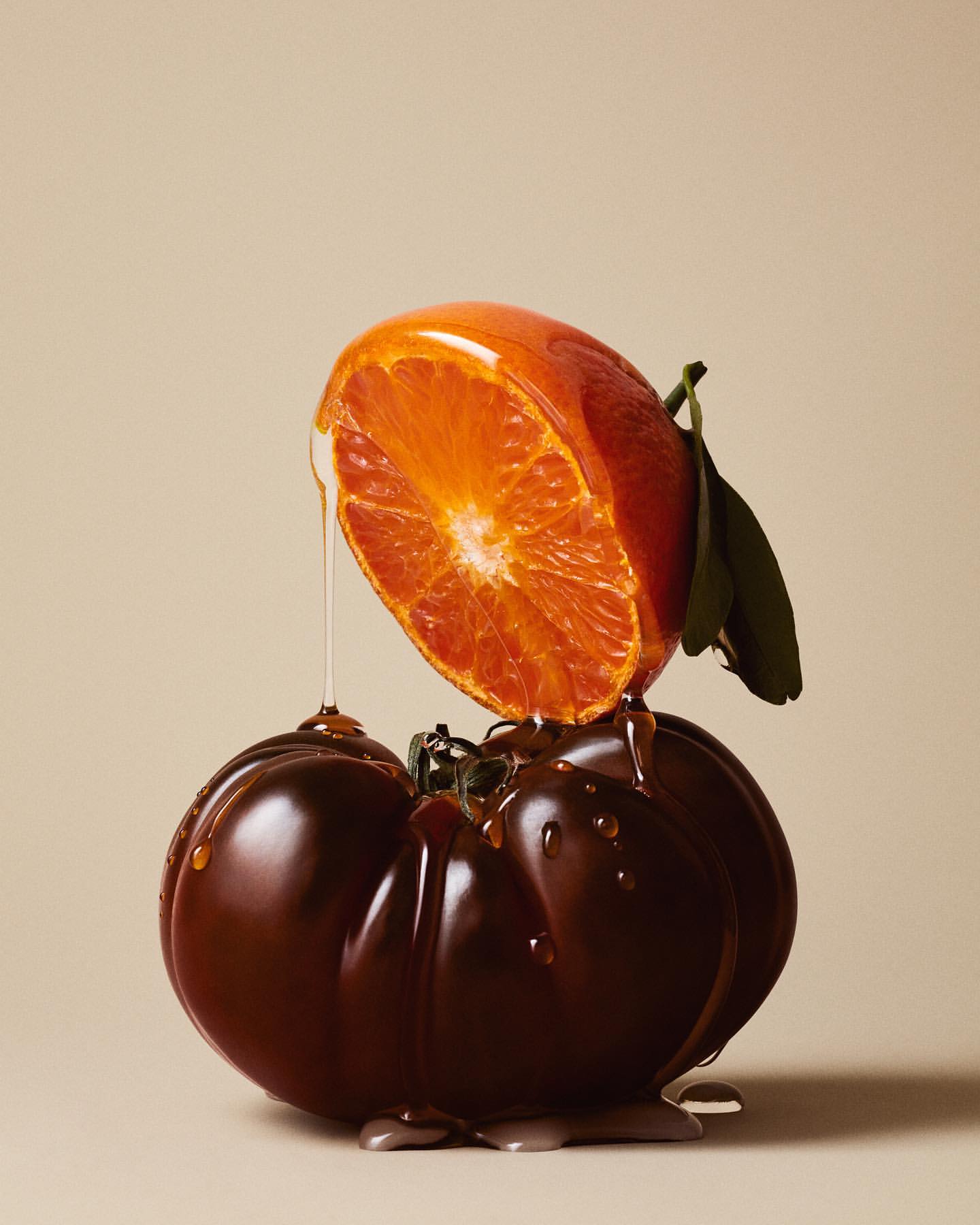 Still life shot of a halved orange resting atop a dark brown tomato, both glazed with honey, set against a neutral-toned background.