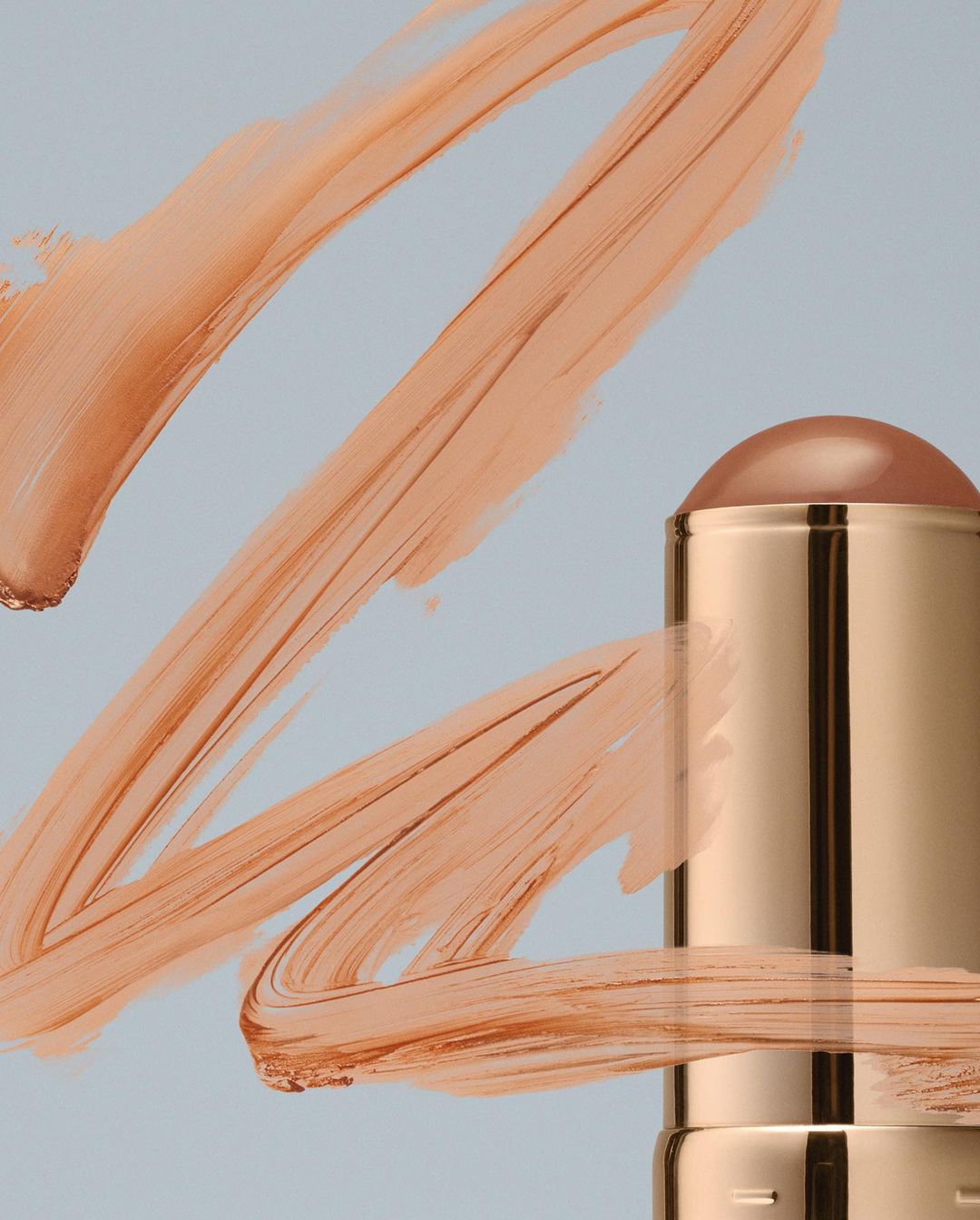 Beauty product shot: Close-up of a tan-colored makeup stick surrounded by tan cream smears on a light blue surface, studio lighting.
