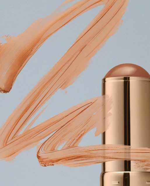 Beauty product shot: Close-up of a tan-colored makeup stick surrounded by tan cream smears on a light blue surface, studio lighting.