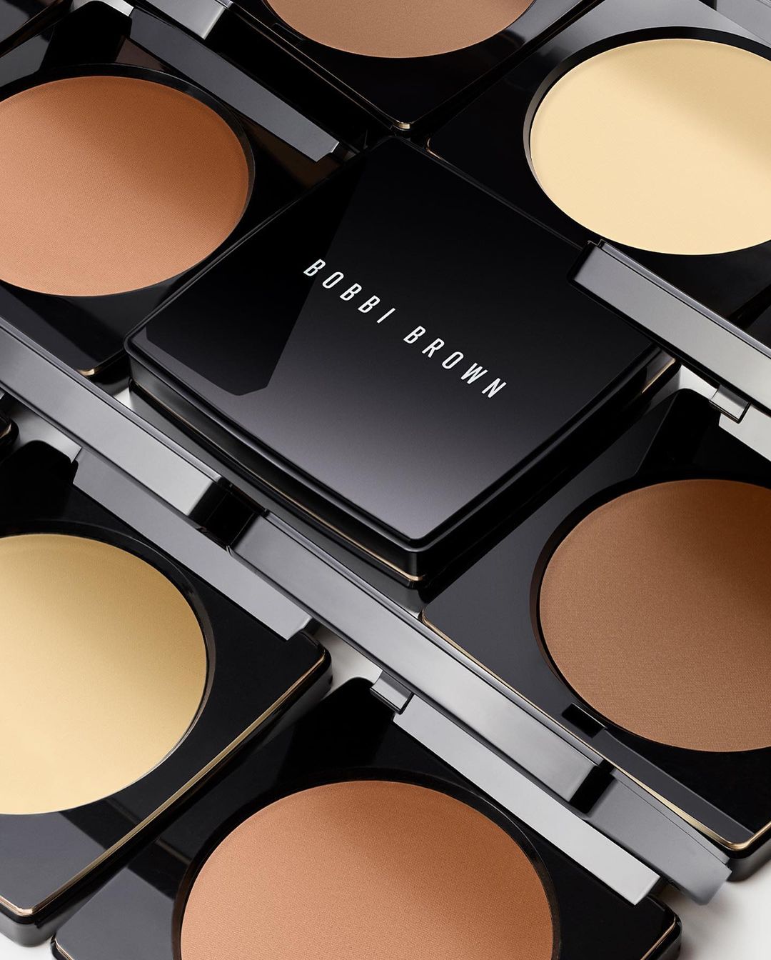 Arrangement of Bobbi Brown compacts in black packaging containing pressed powder in a range of skin tone shades.