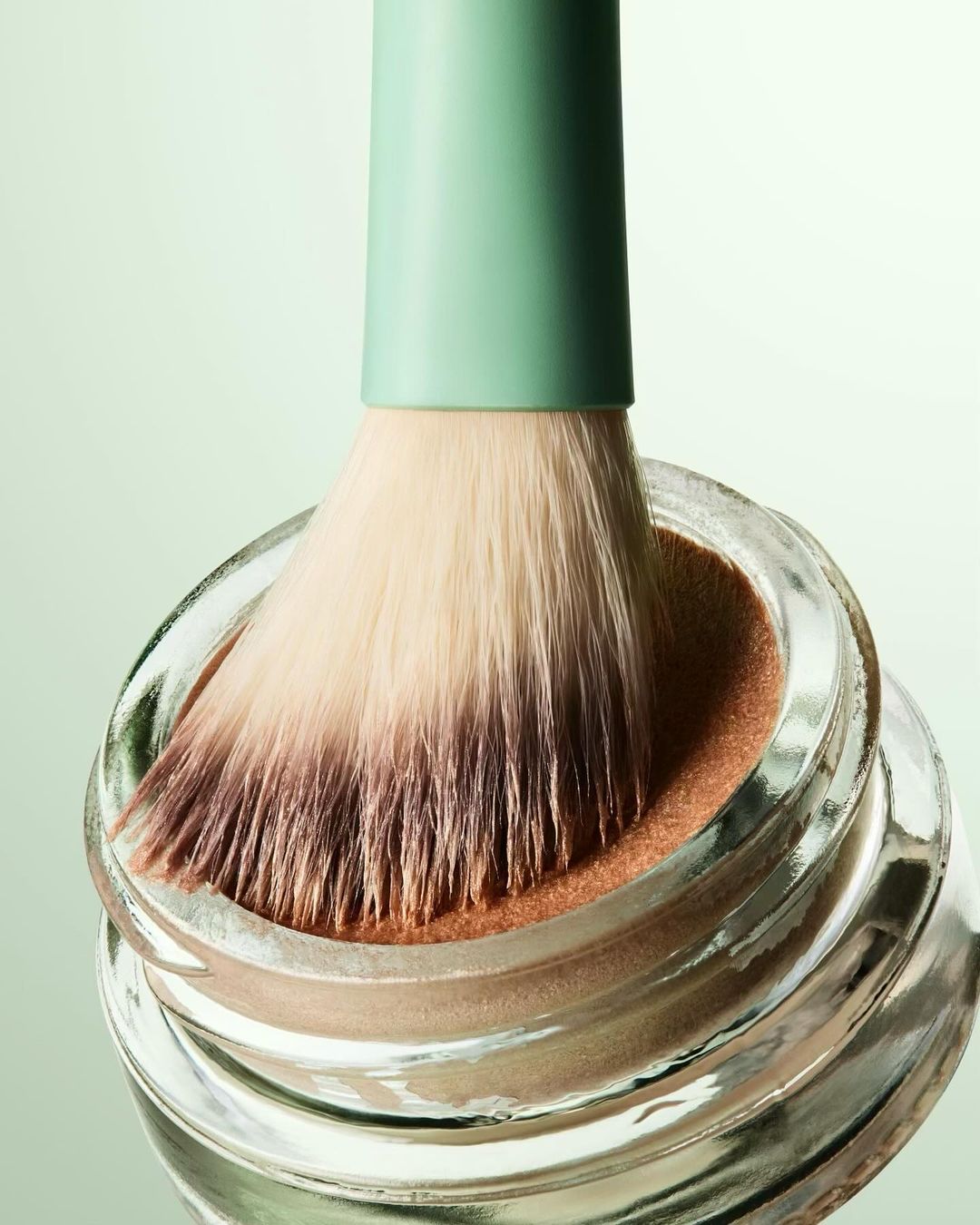 Close-up of a cosmetic powder brush dipped in a clear jar filled with light brown powder, set against a pale green background.