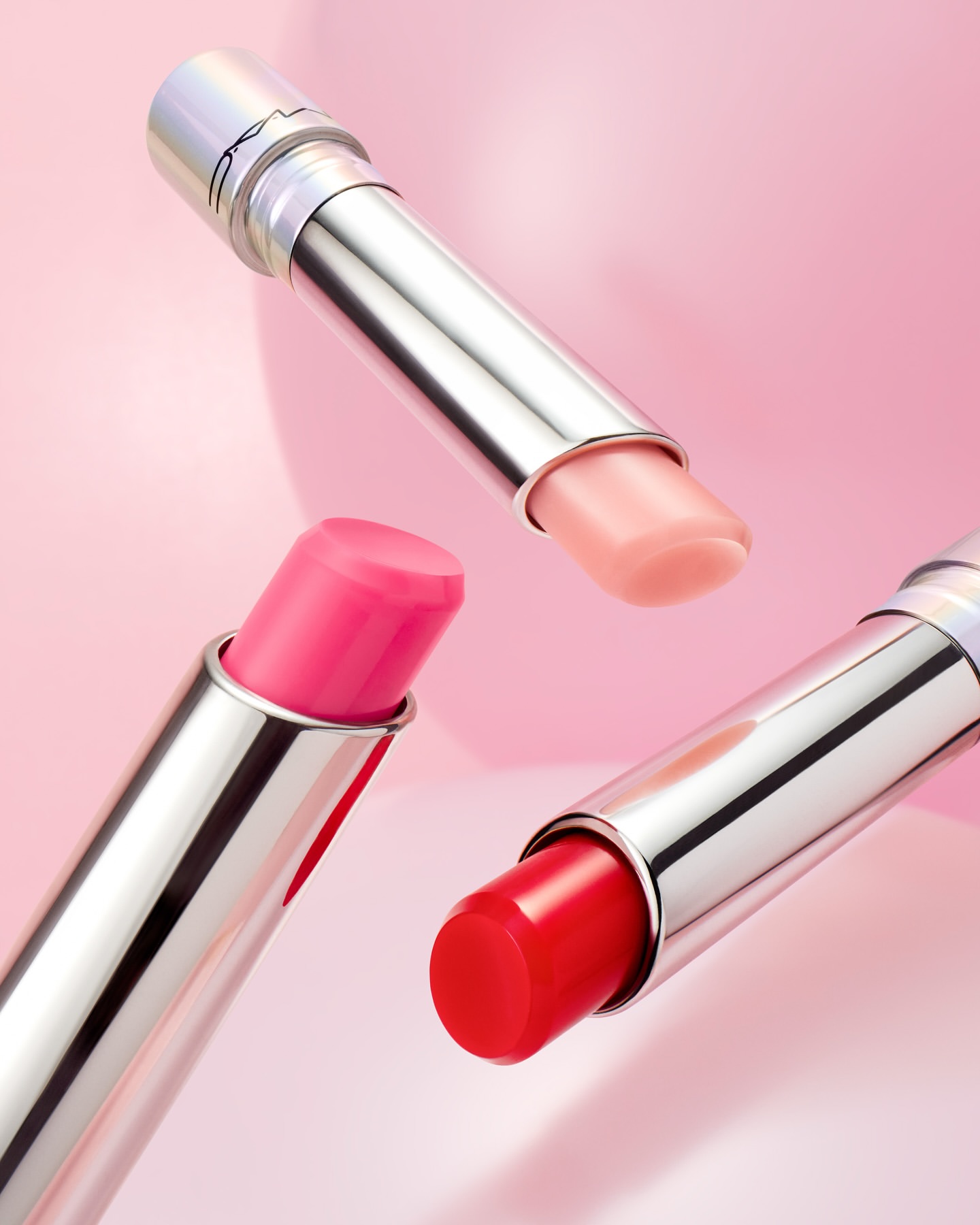 Three lipsticks with silver tubes and pink, red, and peach shades floating in front of a blurred pink background in a beauty still life.