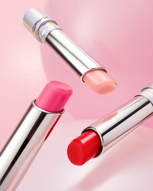 Three lipsticks with silver tubes and pink, red, and peach shades floating in front of a blurred pink background in a beauty still life.