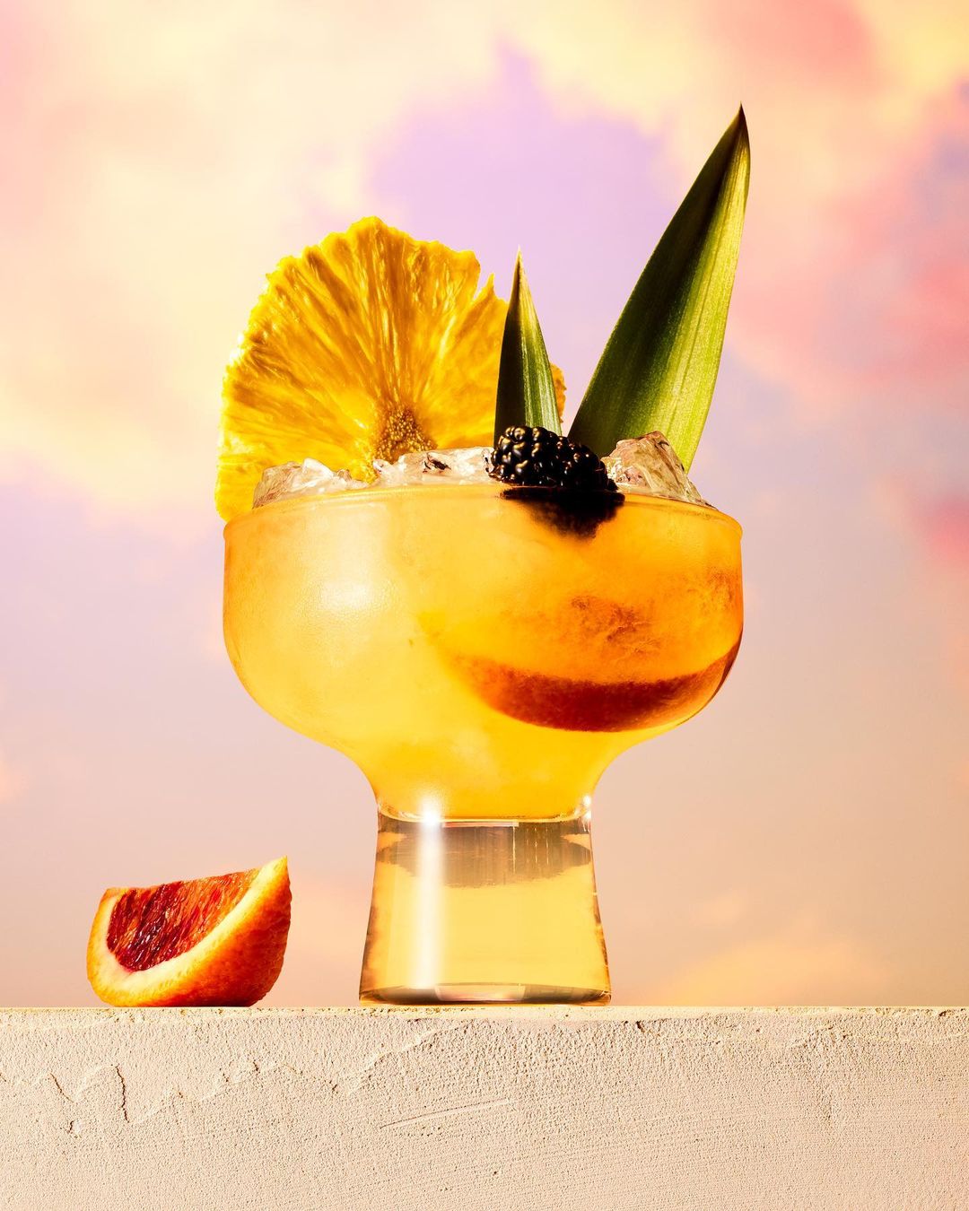 A vibrant cocktail in a stemmed glass with tropical fruit garnishes, including pineapple and blackberry, set against a soft sky background.