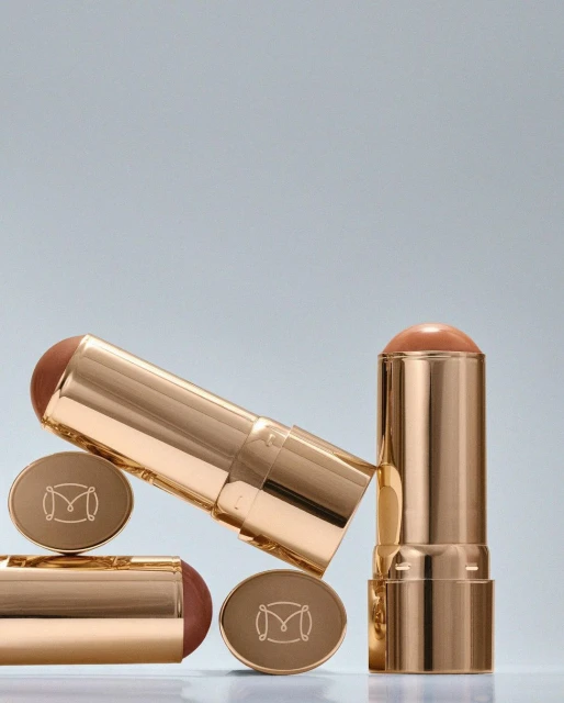 Three gold-toned makeup sticks with brown-colored product, arranged against a gradient blue background, product photography.