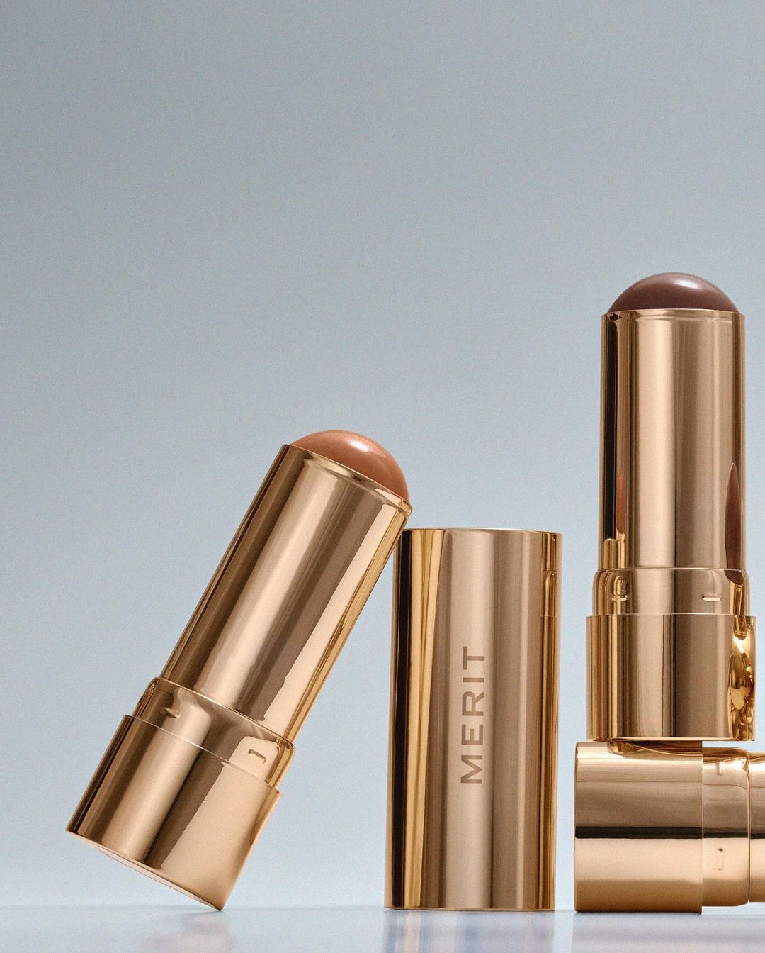 Product shot of three Merit brand makeup sticks in gold tubes with tan and brown product against a gradient grey background.