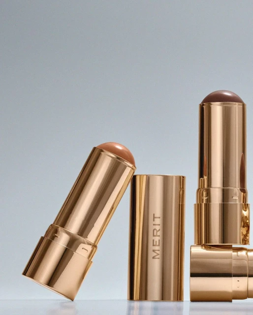 Product shot of three Merit brand makeup sticks in gold tubes with tan and brown product against a gradient grey background.