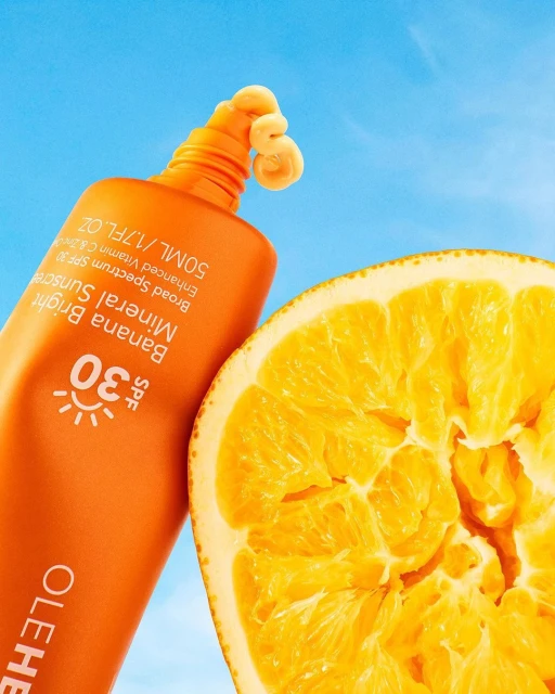 Ole Henriksen Banana Bright Mineral Sunscreen SPF 30 tube next to a sliced orange against a blue sky backdrop.