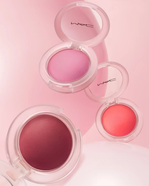 Beauty still life of MAC cosmetics blush compacts in shades of pink, peach, and rose with a soft pink background and diffused lighting.