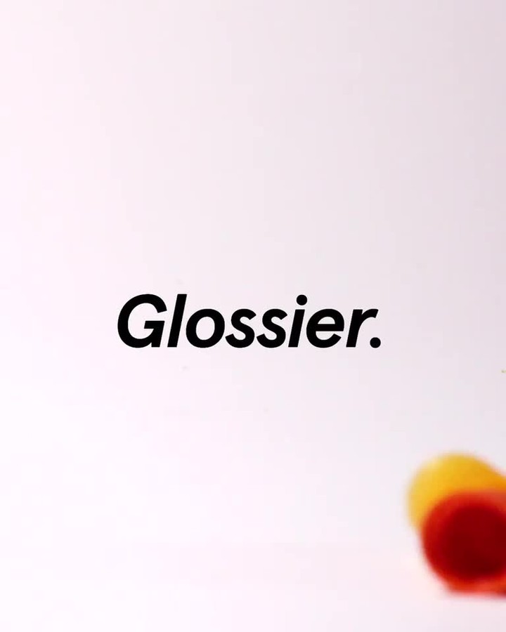 Glossier brand logo and a container with red and yellow contents against a white background in a close-up studio photograph.
