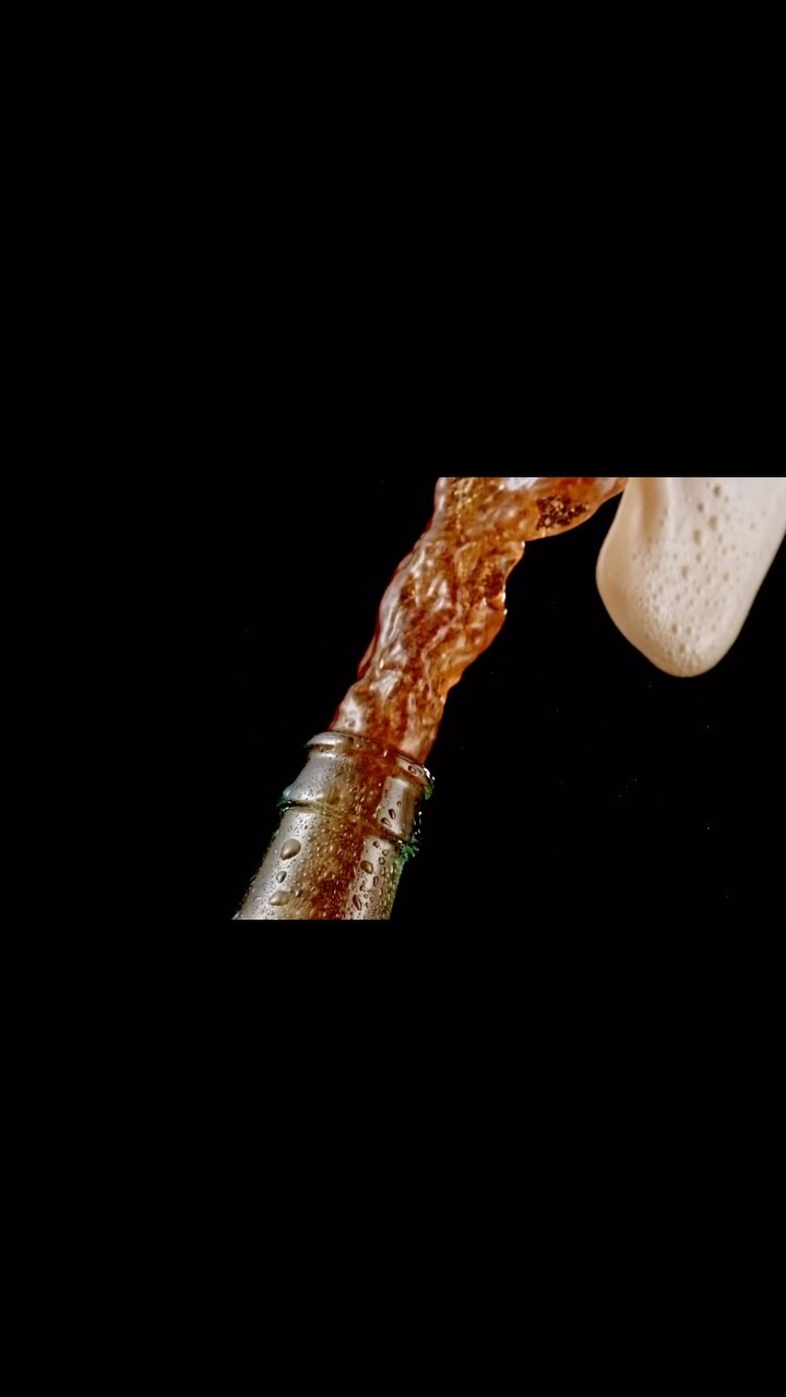 Brown liquid pours from a bottle with condensation and foam on a black background, suggesting a beverage advertisement.