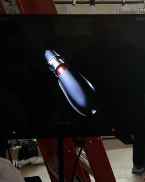 A monitor displaying a digitally rendered futuristic device with a sleek design against a dark background. Display is mounted on a stand.