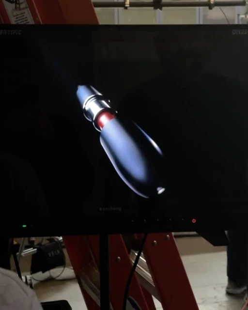 A monitor displaying a digitally rendered futuristic device with a sleek design against a dark background. Display is mounted on a stand.