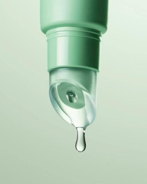 Close-up of a green tube dispenser with a clear liquid and a single water droplet falling in a light green studio backdrop.