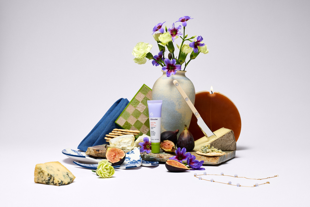 Still life composition featuring skincare product by Glossier, an assortment of gourmet cheeses, crackers, fresh figs, and vibrant flowers.