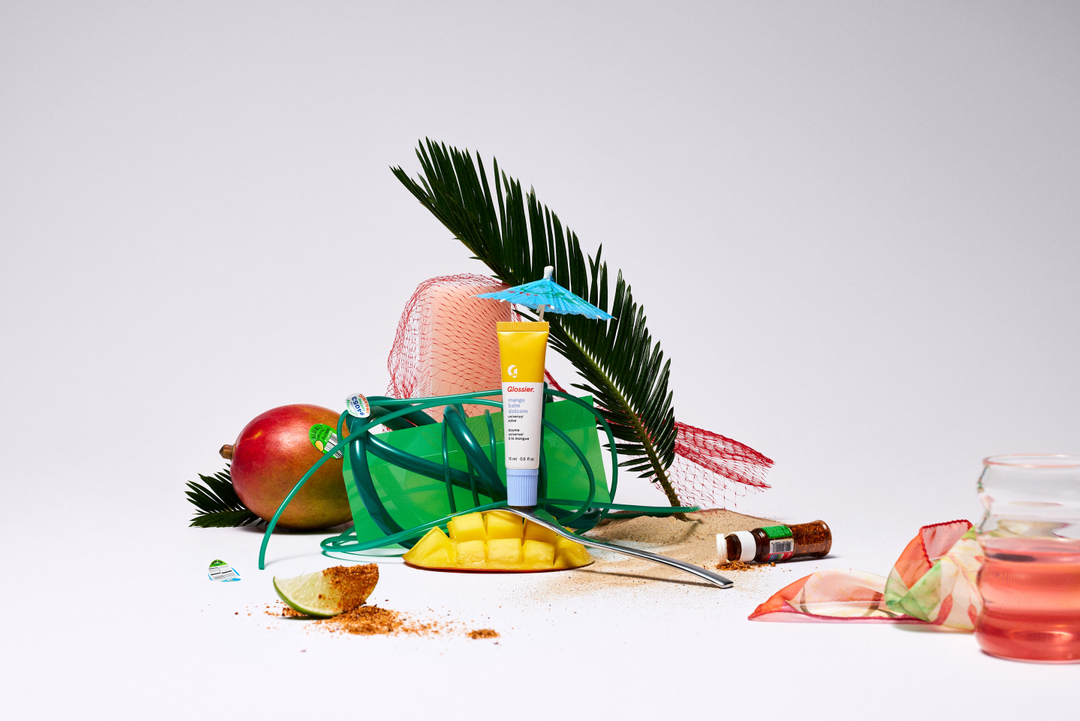 Still life featuring Glossier skincare tube with fruit like mango and lime, green bag, palm leaf, paper umbrella, and pink liquid.