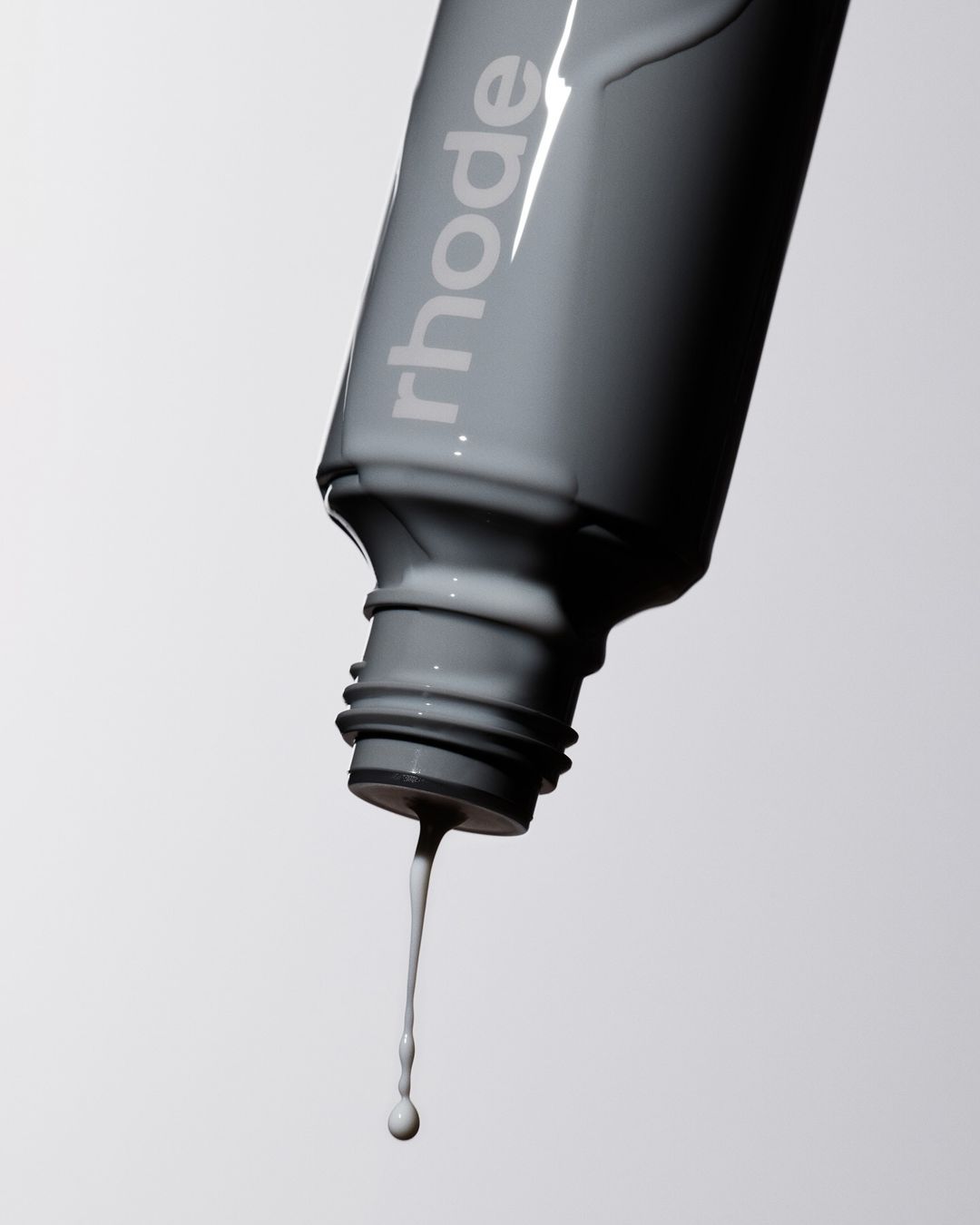Close-up of gray Rhode skincare bottle with viscous liquid dripping from the open cap against a light background in a beauty studio.
