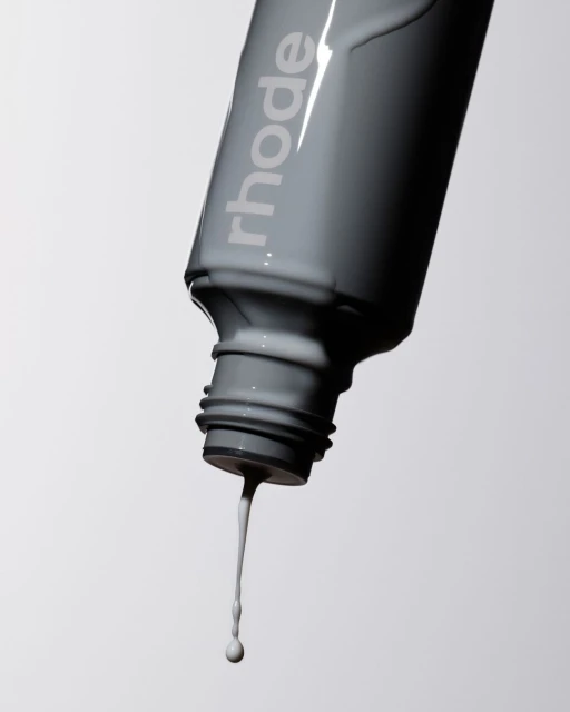 Close-up of gray Rhode skincare bottle with viscous liquid dripping from the open cap against a light background in a beauty studio.
