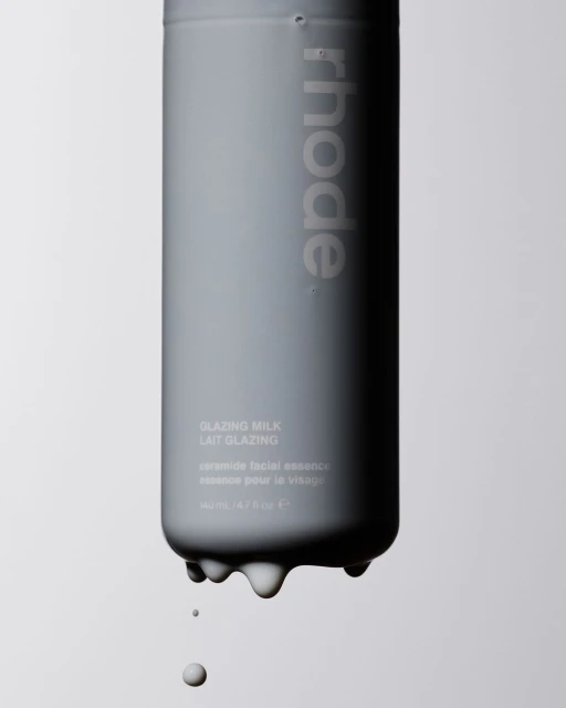 Gray cosmetic bottle of Rhode Glazing Milk with liquid dripping from the bottom, against a light gray background in a studio setting.