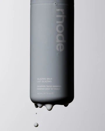 Gray cosmetic bottle of Rhode Glazing Milk with liquid dripping from the bottom, against a light gray background in a studio setting.