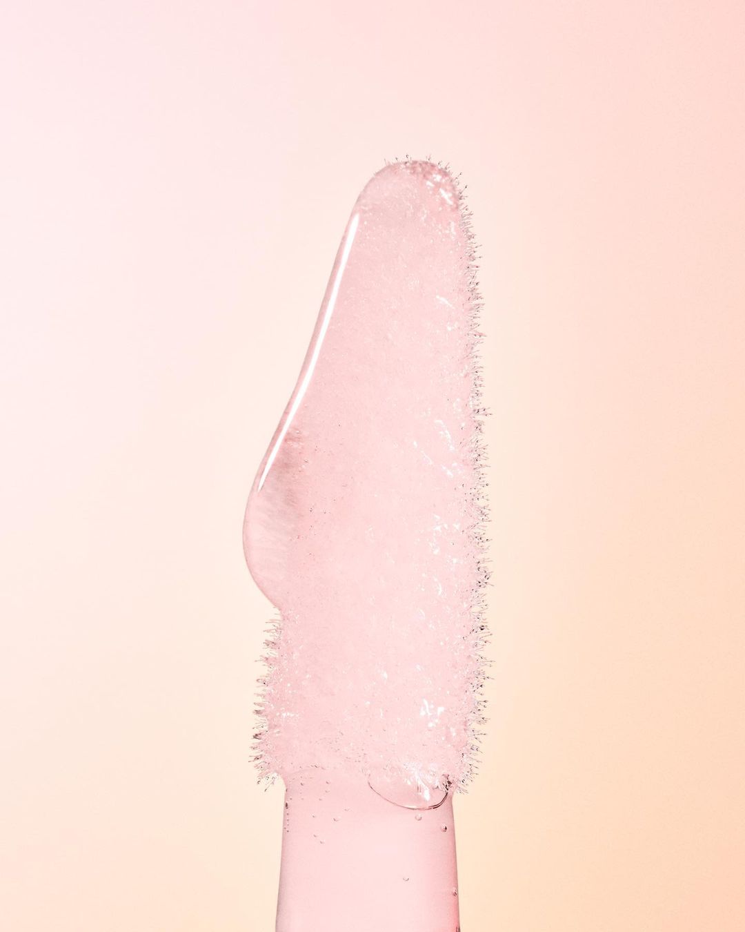 Close-up of a lip gloss applicator with a soft pink hue, showing the fine bristles and texture of the product against a light background.