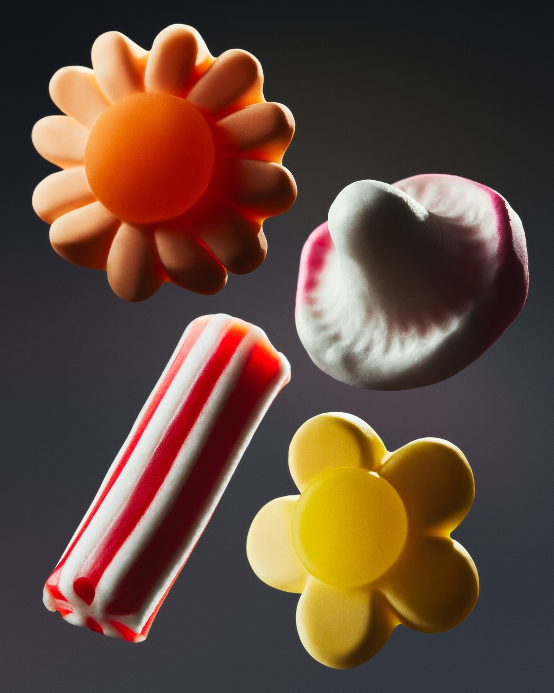 An overhead, close-up studio shot shows four brightly colored gummy candies in flower and striped-stick shapes against a gray background.