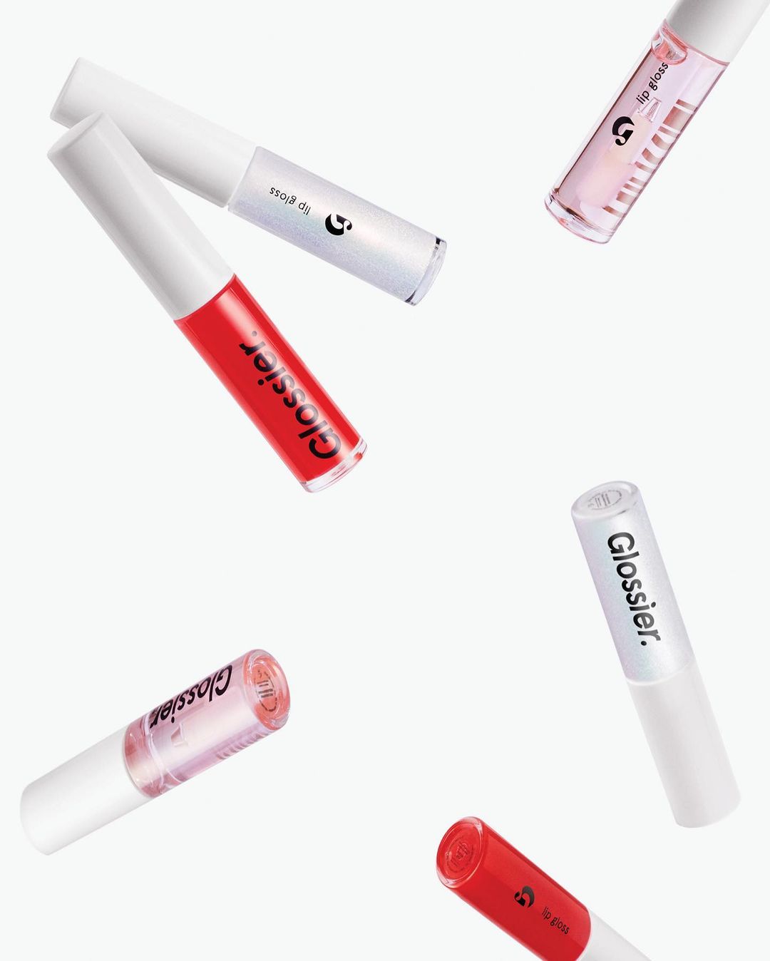 Five Glossier brand lip glosses in a variety of colors with white caps floating on a bright white background in an eye-level product shot.