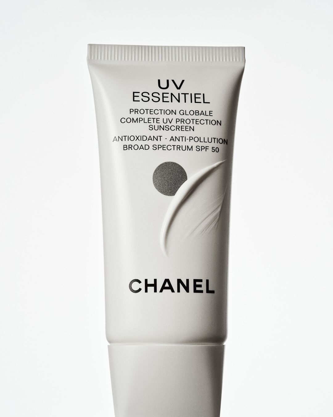 Chanel UV Essentiel sunscreen tube on a white background. A white tube with black lettering is centered in a vertical format.
