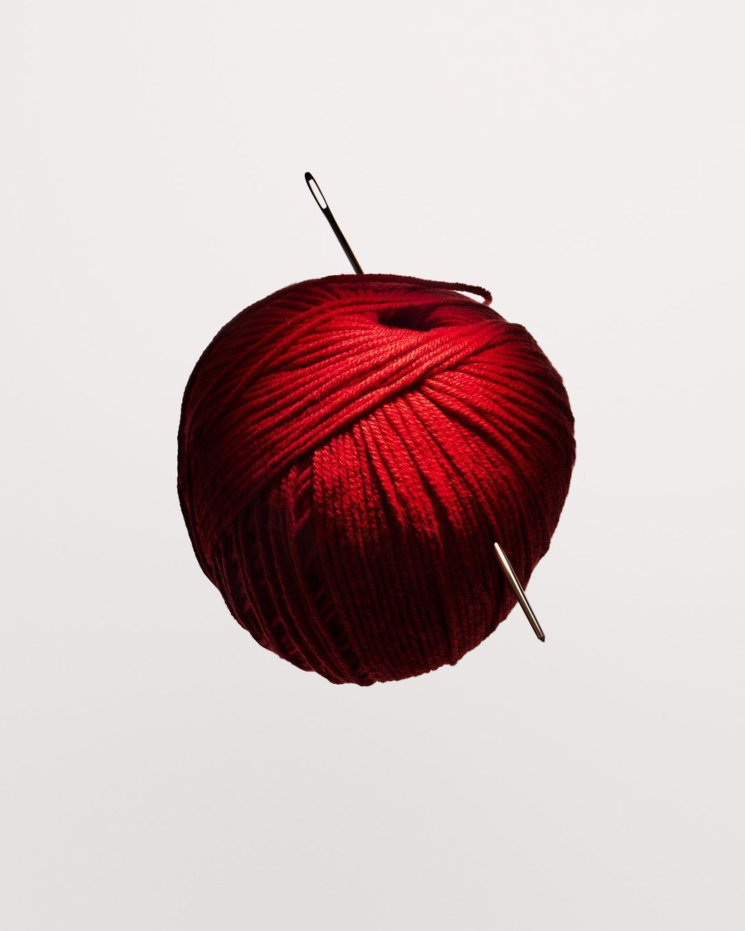 Red yarn ball with sewing needles on a white background in a studio shot, showcasing texture and crafting materials.