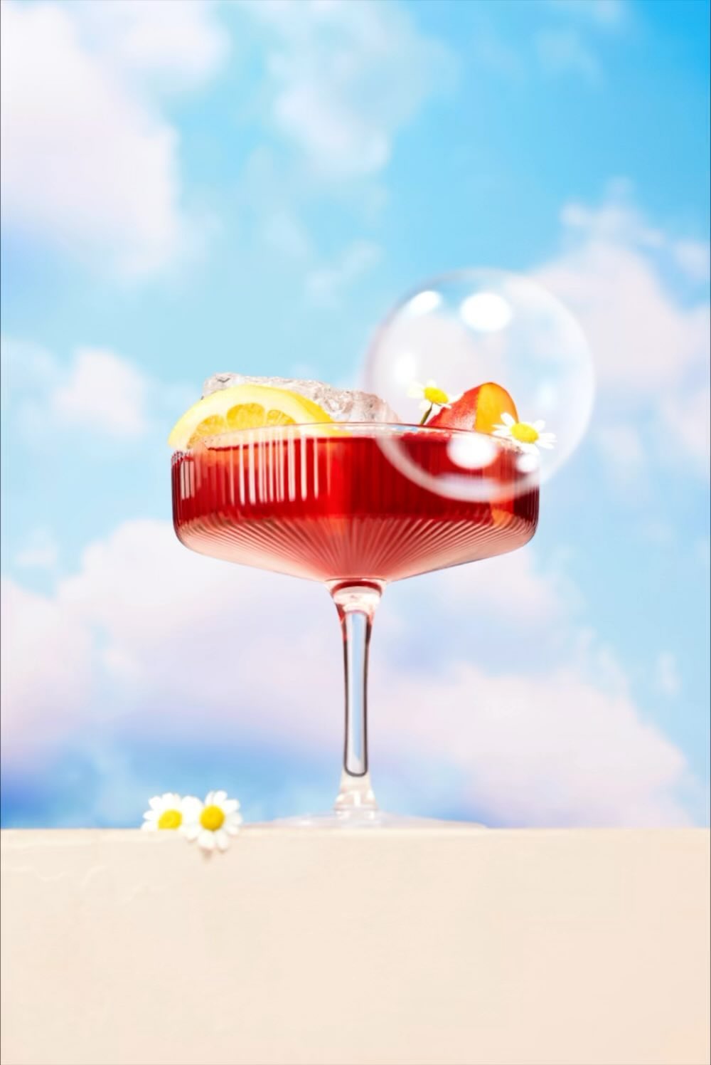Red cocktail drink with fruit, ice, and decorative flowers served on a table against a cloudy sky background in a studio setting.